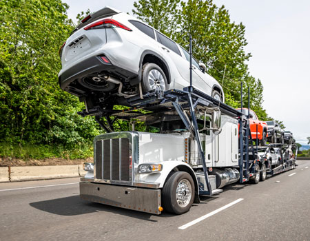 White industrial-grade open car hauler driving on a wide highway with trees alongside, offering expedited car transport services with specialized hydraulic trailer